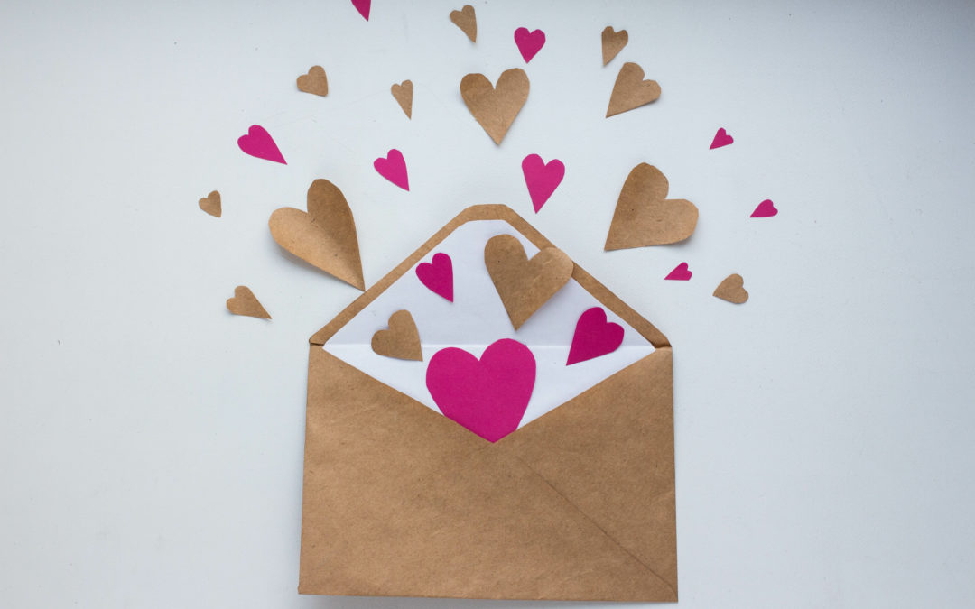 Image of an open brown greeting card envelope with pink, red, and brown paper hearts floating out of it.