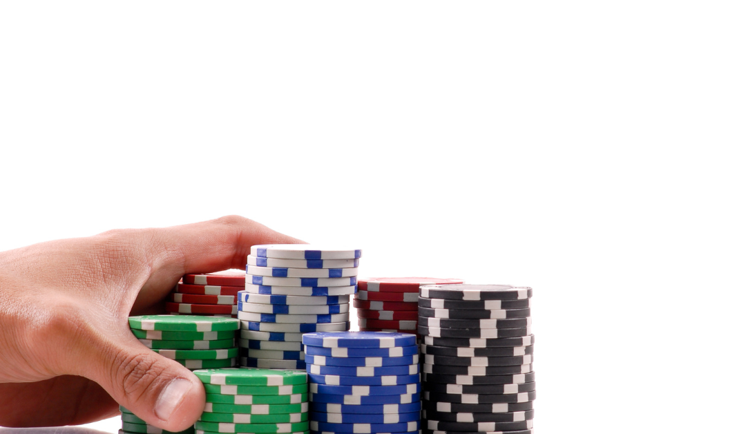 Image of hand pushing, red, white, blue, green, and black poker chips across a table from the left hand side.