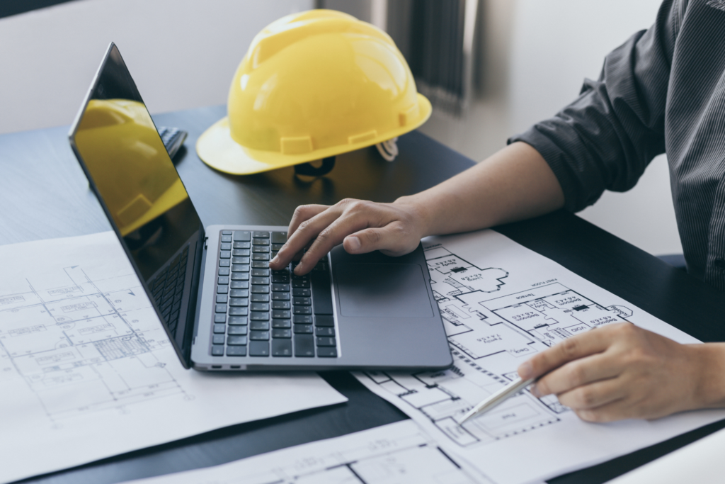 Construction professional using a laptop while reviewing building plans with a yellow hard hat nearby, illustrating business planning and project management in the construction industry.